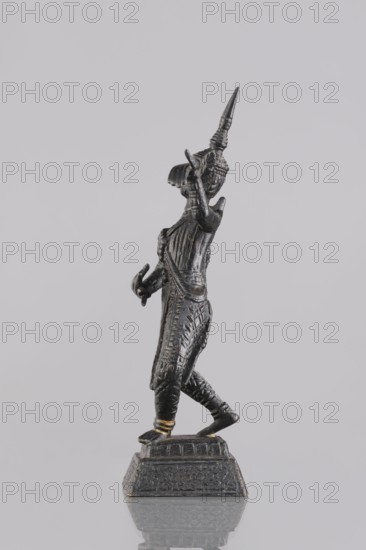 Dancer figurine, Southeast Asian art, musicians, Southeast Asia culture, 1st half of the 20th century contemporary product, Cambodia, figurine, sculpture, height 12.7 cm, A dark metallic figurine of a standing person with a pointed headpiece is shown in profile against a light gray background, with detailed textured clothing and raised arm gestures, a few small gold-colored accents on the ankles and base, and a patterned square pedestal beneath the figure.