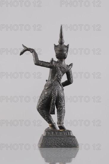 Dancer figurine, Southeast Asian art, musicians, Southeast Asia culture, 1st half of the 20th century contemporary product, Cambodia, figurine, sculpture, height 12.7 cm, A small dark metallic statue viewed from behind showing a standing figure on a decorated rectangular base, the figure wearing an ornate pointed headdress and richly textured clothing with patterned trousers and a draped sash, one arm raised with a curved hand gesture and the other arm bent at the waist, the surface showing highlights of lighter metallic tones against the darker metal, all set against a pale neutral background with a subtle reflection of the base below.