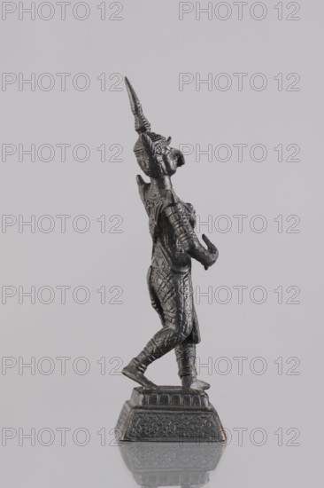 Dancer figurine, Southeast Asian art, musicians, Southeast Asia culture, 1st half of the 20th century contemporary product, Cambodia, figurine sculpture, height 12.7 cm, A small dark metallic statue of a standing human figure on a decorative square base, shown in profile facing right, wearing a tall pointed headdress and textured clothing with ornate patterns, with one arm bent forward and the other slightly raised, visible detailing on the hair, face, and costume, and colors including shades of dark gray and black against a light gray background.