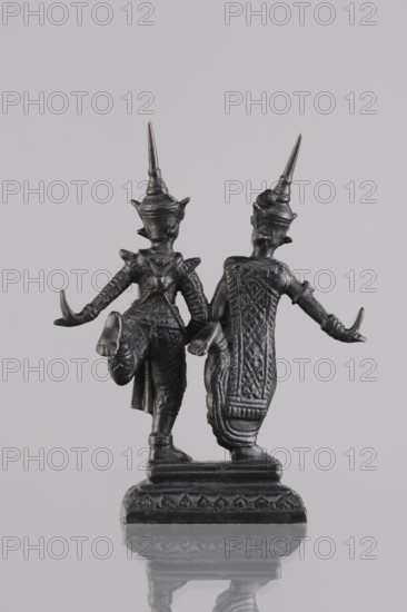 Figurines of a dancing couple, Southeast Asian art, dance, Southeast Asia culture, unknown provenance; figurines of dancers slightly turned to face each other; 1st half of the 20th century contemporary product; Cambodia; figurine, sculpture; height 10.8 cm., A small dark metal statuette of two standing figures joined at the base, shown from the back against a light gray background; the figures wear tall pointed headdresses and have outstretched arms with curved hands, textured garments with patterned details, and one figure has a raised leg, while the rectangular base has decorative motifs and the surface beneath reflects the statuette. The visible colors are dark gray to black on the statuette, light gray in the background, and subtle lighter gray reflections.