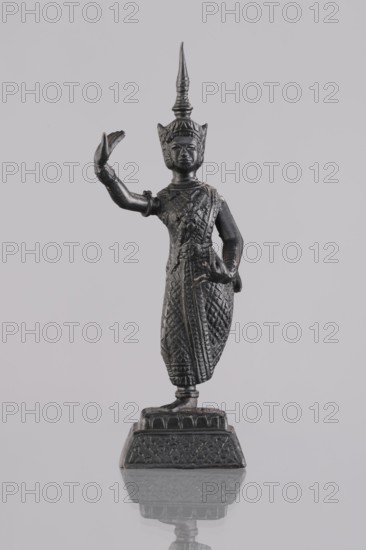 Dancer figurine, Southeast Asian art, Southeast Asia culture, dancers, provenance unknown, 1st half of the 20th century, Cambodia, figurine sculpture, height 12.7 cm, A dark metallic statue of a standing figure is centered against a pale gray background, showing a tall pointed headdress and a detailed textured garment with intricate patterns; one arm is raised with an open hand while the other rests near the waist, and the figure stands on an ornate rectangular base that casts a faint reflection on the smooth surface below. Colors visible include dark gray to black on the statue and various shades of light gray in the background and reflected surface.