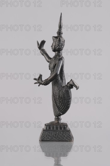 Dancer figurine, creator unknown, Southeast Asian, Southeast Asia culture; dancers; provenance, 1st half of the 20th century; Cambodia; figurine sculpture; height 12.7 cm., A small dark metallic figurine with a smooth reflective finish stands on a patterned pedestal, showing a profile view of a figure wearing an ornate pointed headdress and detailed patterned garments; the figure has one arm raised with an open hand and the other extended forward with splayed fingers, one leg bent at the knee, and a textured skirt with carved decorative lines. The background is a uniform pale gray, and the surface beneath the pedestal reflects the base and lower part of the figurine. Colors visible include dark metallic tones on the figurine and pedestal, pale gray in the background, and subtle lighter gray in the reflection.