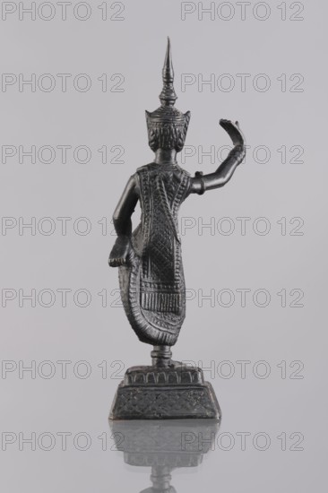 Dancer figurine, creator unknown, Southeast Asian art, Southeast Asia culture, dancers, provenance unknown, 1st half of the 20th century contemporary product, Cambodia place of origin, figurine, sculpture, height 12.7 cm, A small dark metal figurine seen from behind standing on a decorative base against a light gray background, showing a tall pointed headdress and detailed textured clothing with patterned relief, one arm bent upward and the other bent downward holding a curved element, with visible highlights and shadows in black and dark gray tones and a subtle reflection of the base on the surface below.