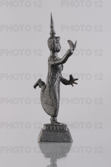Dancer figurine, Southeast Asian art, Southeast Asia culture, dancers, 1st half of the 20th century, Cambodia, figurine, sculpture, height 12.7 cm, A dark metallic statue of a standing figure with a tall pointed headdress and an elongated ornate body, shown in profile facing right; the figure has one arm raised with an open hand and the other arm extended forward with fingers splayed, and the legs are bent with one foot lifted behind; intricate carved patterns cover the figure's clothing and headdress; the statue stands on a patterned square base that is reflected on the glossy surface beneath; visible colors include dark gray, black, and subtle highlights of silver against a light gray background.