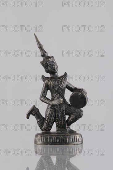 Musician figurine, Southeast Asian art, Southeast Asia culture, musicians, 1st half of the 20th century contemporary product, Cambodia place of origin, sculpture, figurine, height 10.8 cm, A small dark metal figurine with a pointed tall headdress is shown kneeling on a round base; the figure wears detailed patterned clothing with raised decorative elements and holds a round object against its side while one hand rests on its torso. The surface of the figurine has subtle highlights of a lighter metallic color against the predominantly dark tone, and the base shows similar highlights. The background is a uniform pale gray, and the figurine and base cast a faint reflection on a glossy surface beneath them.