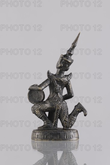 Musician figurine, Southeast Asian art, Southeast Asia culture, musicians, 1st half of the 20th century contemporary product, Cambodia, sculpture, figurine, height 10.8 cm, A small dark metallic figurine with traces of gold paint shows a kneeling figure wearing an ornate pointed headdress and richly patterned clothing; the figure holds a round object near one knee and rests on a circular decorated base, all set against a pale gray background with a reflection visible beneath the base.