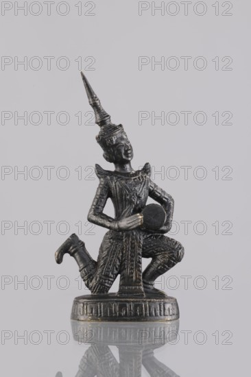 Musician figurine, Southeast Asian art, musicians, Southeast Asia culture, 1st half of the 20th century contemporary product, Cambodia, sculpture, figurine, height 10.9 cm, A small dark metallic statue of a kneeling figure wearing an ornate pointed headdress and detailed patterned clothing is shown against a pale gray background, with the figure holding a round object at chest level and resting on a round base; visible colors include dark gray and hints of gold.