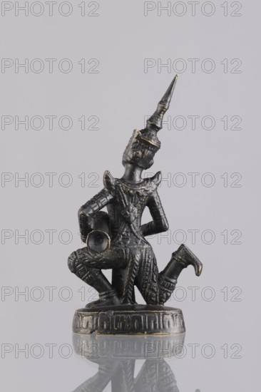 Musician figurine, Southeast Asian art, musicians, Southeast Asia culture, 1st half of the 20th century contemporary product, Cambodia place of origin, sculpture, figurine, height 10.9 cm, A small dark metallic figurine with worn gold highlights shows a kneeling figure wearing an ornate pointed headdress and detailed patterned clothing, holding a cylindrical object against its side; the figure stands on a round decorated base and is positioned against a light gray background with a faint reflection visible beneath it.