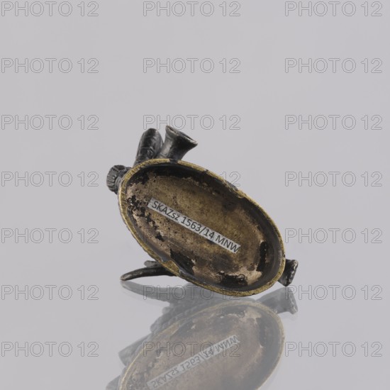 Musician figurine, Southeast Asian art; subject: musicians; culture: Southeast Asia, Cambodia; provenance, 1st half of the 20th century contemporary product; sculpture, figurine; height 10.9 cm, A small metallic object with a worn oval cavity and a raised rim sits tilted on a reflective surface, showing a mirrored reflection beneath. The object has a mix of dark gray or black metal and areas of dull gold or brass along the rim and inner cavity, with patches of darker corrosion or discoloration inside. A few protruding shapes extend from one side of the oval, appearing rounded and tubular in dark gray or black. The background is a pale light gray.