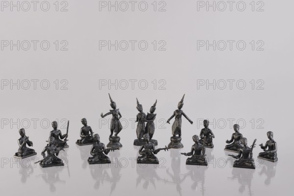 Set of fourteen figurines of musicians, Southeast Asia, art of Southeast Asia, music, dance, musical instruments, 1st half of the 20th century, Cambodia, sculpture, figurine, A group of small dark metallic figurines arranged on a reflective surface against a pale gray background, showing seated figures holding various instrument-like objects and standing figures wearing pointed headdresses with outstretched arms, with visible reflections on the glossy surface and colors including dark gray and black along with the light gray of the background.