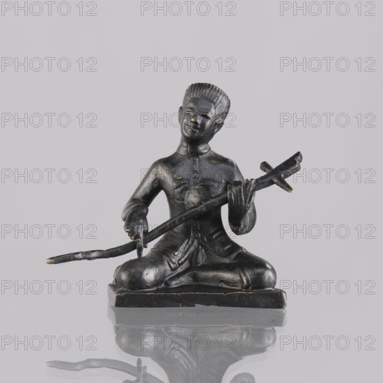 Musician figurine, art of Southeast Asia, Southeast Asia culture, musicians, 1st half of the 20th century contemporary product, Cambodia, sculpture, figurine, height 6.4 cm, A small dark statue of a seated figure shown from the front, positioned on a rectangular base and seated cross-legged, with one arm resting on a bent rod-like object and the other hand holding a longer branch-like object that extends outward; the figure has textured hair combed upward, a faint smile, and clothing with visible folds and buttons; the surface shows small areas of lighter worn spots giving hints of a metallic sheen; the background is a pale neutral gray and the base and figure cast a soft reflection on the glossy surface below.