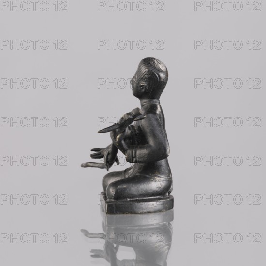 Musician figurine, art of Southeast Asia, Southeast Asia culture, musicians, 1st half of the 20th century contemporary product, Cambodia, sculpture, figurine, height 6.4 cm, A small dark statue of a seated figure shown in profile against a pale gray background, the figure appears to be kneeling on a rounded base and holds an object with several elongated elements in its arms; the surface of the statue is a glossy black with subtle areas of worn lighter highlights and faint hints of gold, and the statue casts a soft reflection on the reflective gray surface beneath it.