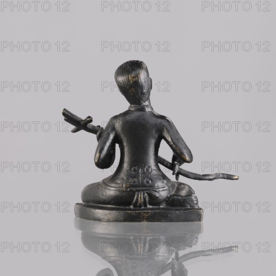 Musician figurine, art of Southeast Asia, Southeast Asia culture, musicians, 1st half of the 20th century contemporary product, Cambodia, sculpture, figurine, height 6.4 cm, A small dark metallic figurine viewed from the back, showing a seated figure with a smooth rounded head and short textured hair, wearing a fitted top and loose lower garment, holding a curved rod-like object across its lap with both hands; the figure sits on a low rounded base and has a few worn lighter patches where the dark surface shows subtle golden highlights, all set against a pale gray background with a faint reflection below.