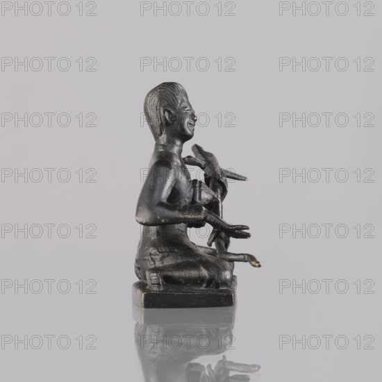 Musician figurine, art of Southeast Asia, Southeast Asia culture, musicians, 1st half of the 20th century contemporary product, Cambodia place of origin, sculpture, figurine, height 6.4 cm, A small dark bronze statuette of a seated human figure on a square base, shown in profile facing right, with textured hair and a visible smile; the figure holds an object with several elongated elements in its hands and arms, and there are subtle worn highlights of a lighter metallic tone on raised areas; the background and surface are light gray and the base casts a faint reflection on the glossy surface beneath.