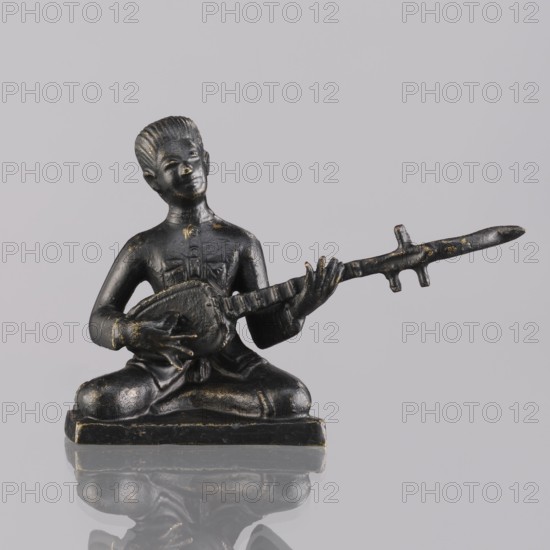 Musician figurine by, Southeast Asia culture, Southeast Asian art; subject: musicians; provenance, 1st half of the 20th century contemporary product; Cambodia place of origin; sculpture, figurine; height 6.2 cm, A small dark statue of a seated person holding a long stringed instrument, shown from the front against a pale gray background, with a reflective surface beneath creating a mirrored reflection; the figure appears in black with subtle gold wear on raised areas, the instrument and base share the same black and gold tones, and the background and reflection include shades of gray.