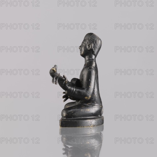 Musician figurine, Southeast Asia culture, Southeast Asian art, musicians, 1st half of the 20th century contemporary product, Cambodia, sculpture, figurine, height 6.2 cm, A small dark figurine sits on a round base with a reflective surface beneath, showing a faint mirrored outline; the figure is seated with legs folded and holds an object in its hands, with a profile view of the head turned to the side; the surface of the figurine is primarily black with several small worn spots revealing a metallic gold tone, and the background is a uniform pale gray.
