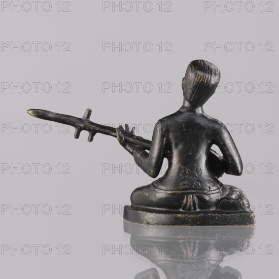 Musician figurine, Southeast Asia culture, Southeast Asian art, musicians, 1st half of the 20th century contemporary product, Cambodia place of origin, sculpture, figurine, height 6.2 cm, A small dark statue of a seated figure viewed from the back, with a smooth dark surface showing worn golden highlights, the figure sitting cross-legged on a rounded base, holding a long horizontal object with small protrusions in its hands, the figure's hair textured and combed back, the base and figure reflecting faintly on a glossy surface; visible colors include dark black, muted gold highlights, and pale gray in the background and reflection.