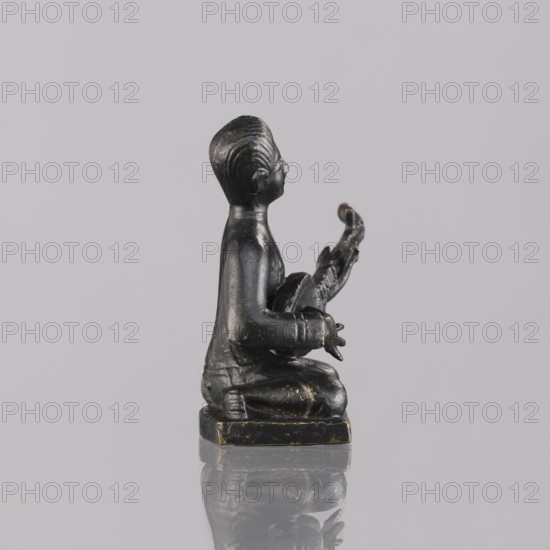 Musician figurine, Southeast Asia, Southeast Asian art, musicians, 1st half of the 20th century contemporary product, Cambodia, sculpture, figurine, height 6.2 cm, A small dark statue of a kneeling human figure is shown in profile against a light gray background, the figure appearing black with subtle gold worn highlights. The figure sits on a square base that shares the same black and gold tones, and it holds a curved object in its hands extending upward. The surface of the statue shows smooth contours and areas of slight wear where the black finish is thin and gold tones peek through, and the base is reflected faintly on the glossy surface beneath it.