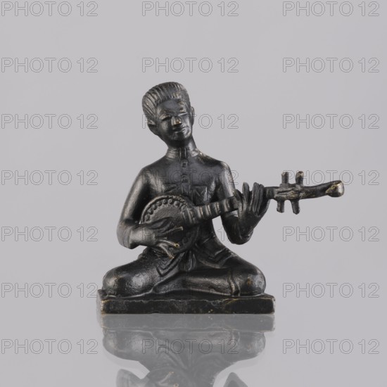 Musician figurine, art of Southeast Asia, Southeast Asia culture, musicians, 1st half of the 20th century contemporary product, Cambodia, sculpture, figurine, height 6.3 cm, A small dark statue of a seated figure holding a stringed instrument; the figure is dark gray to black with subtle worn highlights of lighter goldish tones, sitting cross-legged on a rectangular base that shares the same dark color and worn highlights, with the instrument extending to the figure's right and showing similar dark and goldish tones, all set against a plain light gray background and reflected faintly on a glossy surface below.