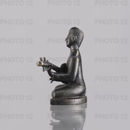 Musician figurine, art of Southeast Asia, Southeast Asia culture, musicians, 1st half of the 20th century contemporary product, Cambodia place of origin, sculpture, figurine, height 6.3 cm, A small dark statue of a seated human figure shown in profile, facing left, with hands extended forward holding a cylindrical object with a few protrusions; the figure has a smooth dark surface with patches of worn lighter tone, sculpted hair texture, and a rounded base that sits on a reflective surface; the background is a uniform pale gray, and the visible colors are dark black, muted gold or bronze wear, pale gray, and reflections of those tones.