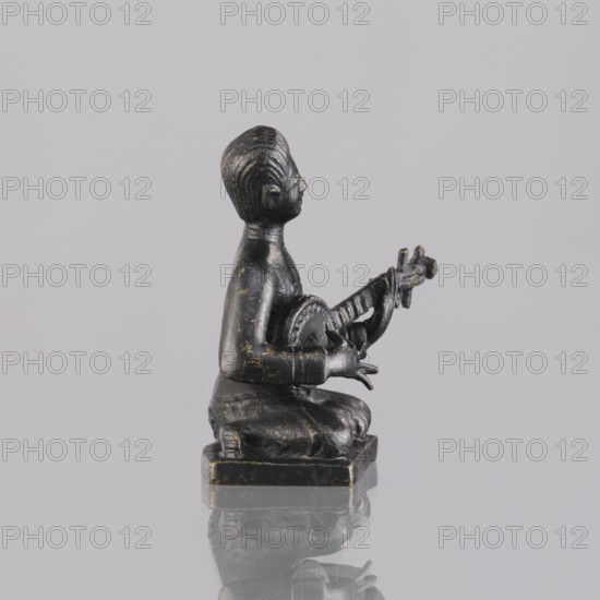 Musician figurine, art of Southeast Asia, Southeast Asia culture, musicians, 1st half of the 20th century contemporary product, Cambodia, sculpture, figurine, height 6.3 cm, A small dark figurine with a worn finish showing flecks of gold sits on a square base, depicting a person kneeling and holding a stringed instrument; the figure has a smooth rounded head with carved hairlines, a defined profile, bent arms with detailed hands positioned on the instrument, a rounded torso, and folded legs with visible drapery folds; the instrument has a circular body and a neck with tuning pegs; the surface colors visible are dark gray to black with gold flecks, and the background is a light gray.