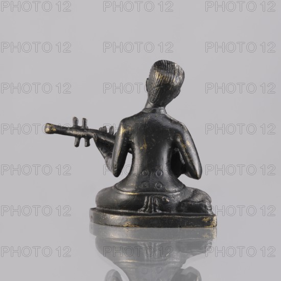 Musician figurine, art of Southeast Asia, Southeast Asia culture, musicians, 1st half of the 20th century contemporary product, Cambodia, sculpture, figurine, height 6.3 cm, A small dark statue viewed from behind showing a seated figure holding a long instrument, with visible worn areas revealing a muted gold tone against the predominant black surface; the figure sits on a rounded base that reflects on the glossy surface below, and the background is a pale gray.