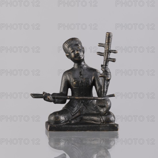 Musician figurine, Southeast Asian art; subject: musicians; culture: Southeast Asia, Cambodia; provenance, 1st half of the 20th century contemporary product; sculpture, figurine; height 6.3 cm, A small dark statue of a seated figure with a shiny surface and worn golden highlights, posed cross-legged on a rectangular base, holding a long horizontal rod with both hands and a vertical staff with several protrusions in one hand; the figure has a sculpted hairstyle and clothing details, and the whole object sits on a reflective pale gray surface against a pale gray background with visible reflections.