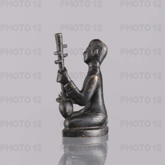 Musician figurine, Southeast Asian art, musicians, Southeast Asia culture, 1st half of the 20th century contemporary product, Cambodia, sculpture, figurine, height 6.3 cm, A small dark figurine sits on a rounded base, shown in profile facing right, with a smooth dark surface accented by worn golden highlights; the figure is seated with legs folded beneath, wearing a long-sleeved garment, and holding a vertical object with several short protrusions and a bulbous element near its lower end; the head has stylized hair and facial features with a serene expression; the figure and base rest on a reflective surface that mirrors their shapes against a pale gray background.