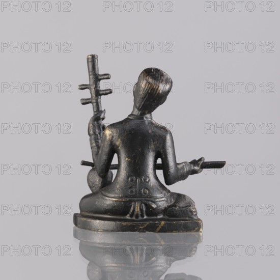 Musician figurine, Southeast Asian art; subject: musicians; culture: Southeast Asia; provenance, date: 1st half of the 20th century contemporary product; place of origin: Cambodia; medium: sculpture, figurine; height: 6.3 cm, A small dark metal figurine is shown from behind, seated on a rounded base with legs crossed and a draped garment; the surface has worn highlights in a muted gold and the metal appears black with subtle brownish tones, with a few scuffed areas revealing brighter metallic flecks. The figure holds a vertical staff with several short horizontal prongs in one hand and a horizontal rod in the other, both matching the same black-and-gold patina. The head is slightly tilted and textured with carved hair striations, and the base sits on a reflective light-gray surface that mirrors the lower portion of the figurine against a plain pale-gray background.
