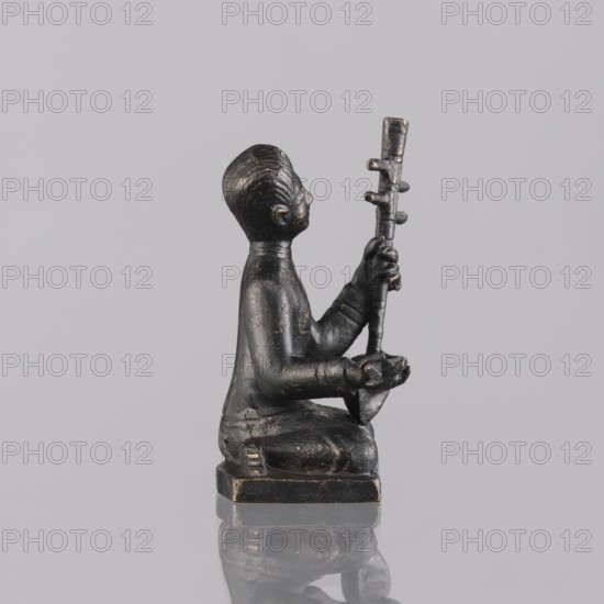 Musician figurine, Southeast Asian art, musicians, Southeast Asia culture, 1st half of the 20th century contemporary product, Cambodia, sculpture, figurine, height 6.3 cm, A small dark figurine of a person kneeling on a square base, shown in profile facing right, with a smooth rounded head and stylized facial features, holding a tall vertical object with a few short protrusions in both hands; the figure and object are mostly black with worn areas revealing a few small patches of lighter, golden tones, set against a plain light gray background with a reflective surface beneath that creates a faint mirrored outline.
