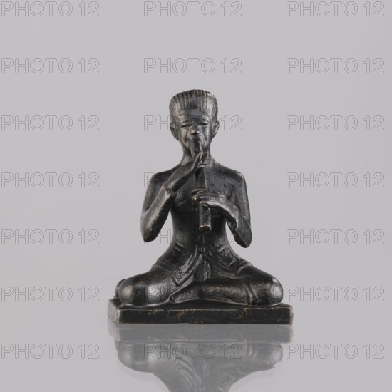 Musician figurine, art of Southeast Asia, Southeast Asia culture, musicians, 1st half of the 20th century contemporary product, Cambodia place of origin, sculpture, figurine, height 6.4 cm, A small dark statue of a seated figure on a rectangular base is shown against a light gray background, the figure sits cross-legged with a straight posture and holds a vertical object to its mouth with both hands, the statue has a textured surface with visible worn highlights in a lighter metallic tone, the figure's hair appears in short ridged rows and the face shows subtle carved features, the clothing and body have carved folds and contours, the base and the statue cast a faint reflection on the glossy surface below, colors visible include dark black, worn metallic gold highlights, light gray in the background, and subtle variations of dark gray on the statue's surface.