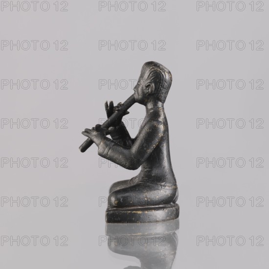 Musician figurine, art of Southeast Asia, Southeast Asia culture, musicians, 1st half of the 20th century contemporary product, Cambodia place of origin, sculpture, figurine, height 6.4 cm, A small dark figurine depicts a person seated on a rounded base, holding a cylindrical object to their mouth with both hands; the figure is shown in profile, with short hair and a simple, smooth garment. The surface of the figurine is primarily black with scattered areas of worn gold or brass-colored highlights, and it rests on a reflective surface that creates a faint mirrored image. The background is a uniform light gray.