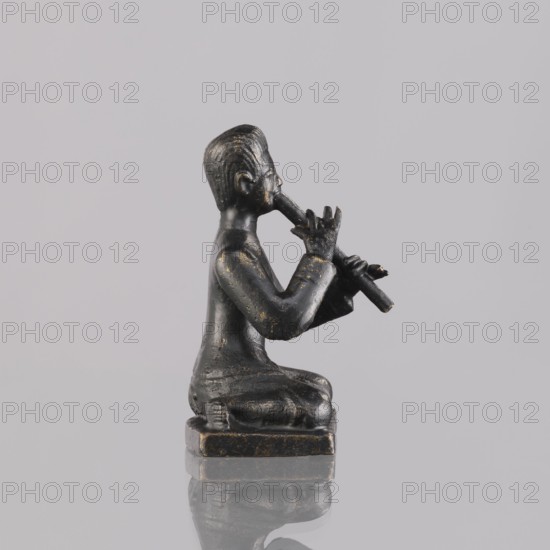 Musician figurine, art of Southeast Asia, Southeast Asia culture, musicians, 1st half of the 20th century contemporary product, Cambodia place of origin, sculpture, figurine, height 6.4 cm, A small dark statue of a seated figure holding a flute to its mouth, rendered in a black material with worn gold highlights, sits on a rectangular base; the figure is shown in profile with short hair, hands positioned on the instrument, and a reflective surface beneath creating a subtle mirror image against a pale gray background.