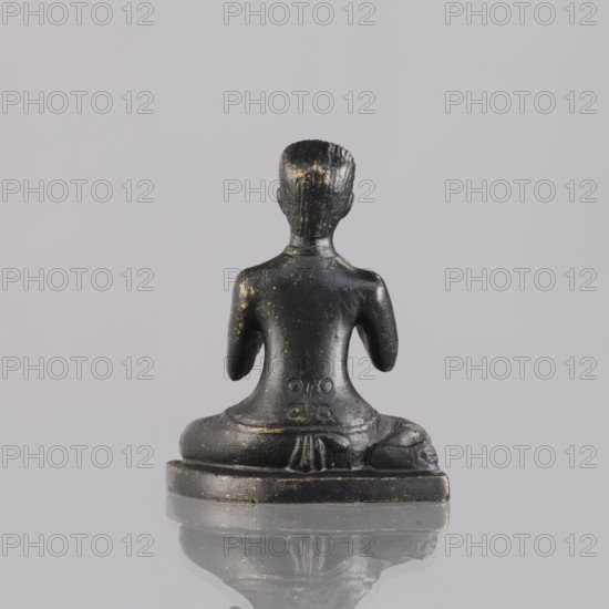 Musician figurine by, art of Southeast Asia, Southeast Asia culture; musicians; unknown provenance; 1st half of the 20th century contemporary product; Cambodia place of origin; sculpture, figurine; height 6.4 cm, A small dark statue viewed from the back showing a seated human figure with a short hairstyle and a smooth back, the surface showing worn areas with gold-colored flecks, the figure seated on a rounded base with draped cloth details at the lower back and hips, set against a flat light gray background with a faint reflection of the base on the surface below; visible colors include black, gold, gray, and a hint of reflection tones.