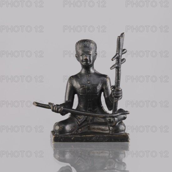 Musician figurine, art of Southeast Asia, Southeast Asia culture, musicians, 1st half of the 20th century contemporary product, Cambodia place of origin, sculpture, figurine, height 6.5 cm, A small dark metallic statuette of a seated figure on a rectangular base, shown against a light gray background and reflected faintly below; the figure sits cross-legged with a straight back and holds a long thin staff with small protrusions in one hand and a curved rod across the lap with the other, the surface showing a worn patina with visible darker black and areas of muted gold accenting, and the figure has stylized hair, defined facial features, and simple clothing details.