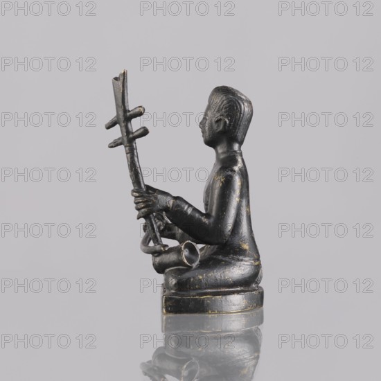 Musician figurine, art of Southeast Asia, Southeast Asia culture, musicians, 1st half of the 20th century contemporary product, Cambodia, sculpture, figurine, height 6.5 cm, A small dark statue of a seated figure is shown in profile holding a tall, vertical object with crosspieces; the figure appears to be seated on a rounded base and is rendered in a dark gray to black material with worn areas showing touches of gold. The figure's hair and facial profile are modeled, and the hands grip the vertical object while a curved element rests near the figure's lap. The background is a smooth pale gray and the surface beneath the statue reflects the object faintly, showing similar dark and gold tones.