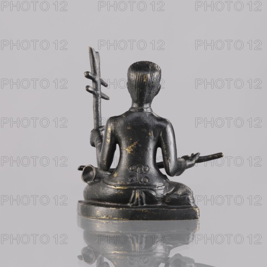 Musician figurine, art of Southeast Asia, Southeast Asia culture, musicians, 1st half of the 20th century contemporary product, Cambodia place of origin, sculpture, figurine, height 6.5 cm, A small dark metal figurine viewed from the back, seated on a rounded base with a smooth reflective surface beneath, showing a few worn areas of lighter metallic color; the figure has short textured hair and a smooth back and shoulders, with one arm raised holding a long slender staff with several short cross pieces and the other arm extended holding a thin rod; there are subtle carved details at the lower back and waist, and the overall colors visible are dark gray to black with touches of goldish wear and the background is a pale muted gray.