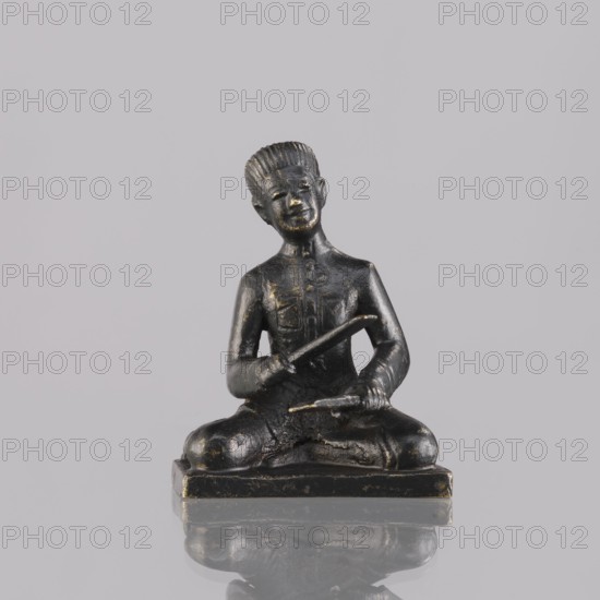 Musician figurine, Southeast Asia culture, musicians, musical instruments, 1st half of the 20th century contemporary product, Cambodia, sculpture, figurine, height 6.4 cm, A small dark statue of a seated figure sitting cross-legged on a rectangular base, rendered in a worn black material with patches of gold showing through; the figure has short ridged hair, a rounded face with a faint smile, and visible ears, and holds a stick-like object in each hand with one held diagonally across the torso and the other resting near the knees; the base and lower portions show texture and wear with the same black and gold tones, and the statue sits on a reflective surface that mirrors its shape against a plain light gray background with shades of gray and pale lavender.