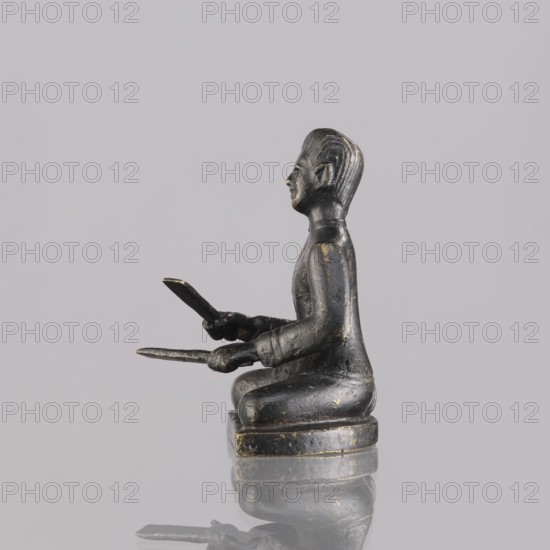 Musician figurine, Southeast Asia culture, musicians, musical instruments, 1st half of the 20th century, Cambodia, sculpture, figurine, height 6.4 cm, A small sculpted figure in a seated pose, shown in profile facing right, appears to be made of a dark material with worn areas revealing a muted gold tone; the figure has stylized hair, a defined face, and is wearing a long-sleeved garment, holding a flat object in each hand with arms extended forward, seated on a low rounded base that reflects faintly on the glossy surface below; the background is a uniform light gray.