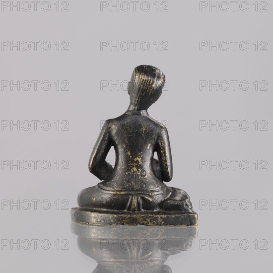 Musician figurine by, Southeast Asia culture; subject: musicians, musical instruments; sculpture, figurine; Cambodia place of origin; 1st half of the 20th century; height 6.4 cm., A small dark statue viewed from behind showing a seated figure on a rounded base, with a smooth back, bent arms resting near the hips, and textured hair; the surface has worn areas with visible gold-colored highlights and scratches on the dark material, and the base reflects faintly on the glossy surface beneath; visible colors include dark black, gold, and soft gray from the background and reflection.