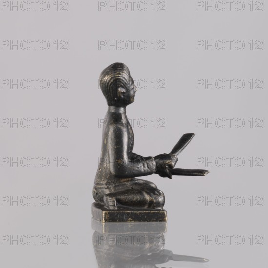 Musician figurine, Southeast Asia culture, musicians, musical instruments, 1st half of the 20th century contemporary product, Cambodia place of origin, sculpture, figurine, height 6.4 cm, A small dark figurine sits on a square base, depicted in profile with a kneeling posture and hands extended forward holding two flat objects; the figure has a smooth, rounded head with carved hair texture, a defined nose and chin, and a simple collar at the neck. The surface shows worn highlights and small patches of lighter metallic color against the predominantly dark material. The base and lower edges also show touches of lighter metallic color where the dark finish is rubbed. The background is a uniform pale gray, and the figurine casts a faint reflection and shadow on the glossy surface beneath it.