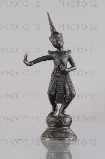 Dancer figurine, dance, Southeast Asian art, Southeast Asia culture, 1st half of the 20th century contemporary product, Cambodia place of origin, sculpture, figurine, height 13.2 cm, A small dark metallic statue of a human figure stands on a round ornate base, the figure wearing a tall pointed headdress and detailed patterned clothing, posed with one arm extended outward and the other bent toward the torso; the surface shows hints of worn metallic highlights. The overall colors visible are dark gray to black with touches of faded gold.