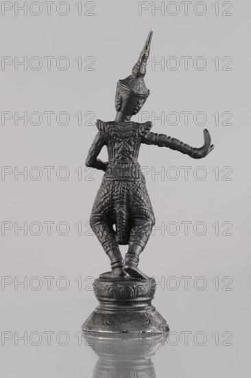Dancer figurine, creator unknown, dancer, Southeast Asian art, Southeast Asia culture, provenance unknown, 1st half of the 20th century contemporary product, Cambodia place of origin, sculpture, figurine, height 13.2 cm, A small dark metal figurine stands on a round stepped base, depicted in a stylized human form wearing an elaborate pointed headdress and ornate patterned clothing; the figure is posed with one arm bent and the other extended outward, weight shifted onto one leg, and the base and lower parts show some lighter worn highlights. The visible colors are dark gray to black with subtle hints of metallic gold and lighter gray where the surface is worn, set against a pale gray background.