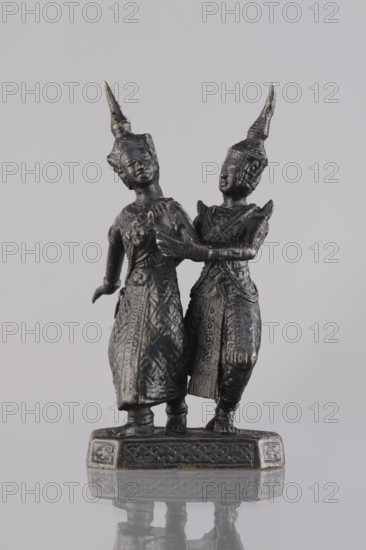 Figurines of a dancing couple, dance, Southeast Asia culture, art of Southeast Asia, 1st half of the 20th century, Cambodia, sculpture, figurine, height 11.7 cm, A small dark metallic statue of two standing figures on a rectangular base, both wearing tall pointed headdresses and long intricately patterned garments; one figure stands slightly forward with an arm extended back while the other stands close behind with an arm touching the first figure’s chest, their faces and hands visible with smooth sculpted features, and the base shows decorative carved patterns; visible colors include dark gray to black metal with subtle hints of bronze or brown tones, and a neutral light gray background.