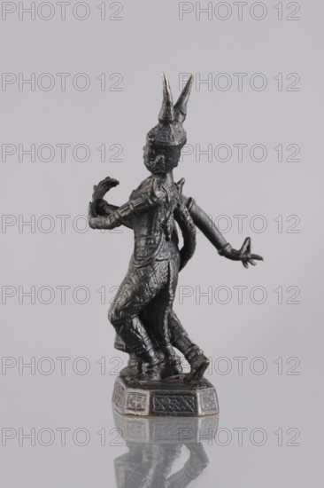Figurines of a dancing couple, dance, Southeast Asia culture, art of Southeast Asia, 1st half of the 20th century, Cambodia, sculpture, figurine, height 11.7 cm, A small dark metallic figurine stands on a decorative base, shown from a rear three-quarter view. The figure has a textured surface with visible tooling marks and wears a long lower garment with folds and patterned details. The figure’s arms are bent and extended in expressive poses, and the head is topped with two pointed protrusions. The base is angular with carved panels and the whole object casts a reflection on the smooth surface beneath. The visible colors are dark gray, black, and a few lighter metallic highlights against a pale gray background.