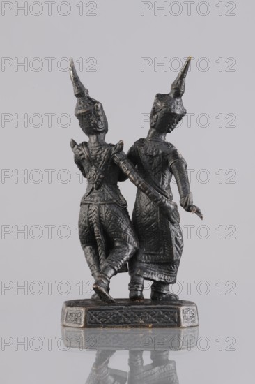 Figurines of a dancing couple, dance, Southeast Asia culture, art of Southeast Asia, 1st half of the 20th century, Cambodia, sculpture, figurine, height 11.7 cm, A small dark metallic figurine of two standing figures joined at the hips on a rectangular base, both wearing tall pointed headdresses and intricately textured clothing with visible patterns and folds; one figure has a raised arm with a bent elbow while the other has an arm extended slightly outward, and both show detailed facial profiles and elongated limbs; the base has carved decorative borders and the surface beneath the base produces a faint mirrored reflection; colors visible include dark gray, black, and hints of metallic bronze.