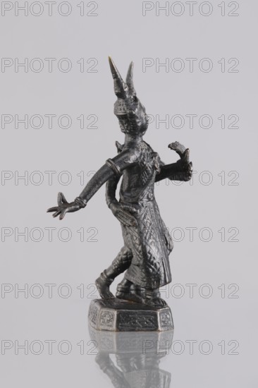 Figurines of a dancing couple, dance, Southeast Asia culture, art of Southeast Asia, 1st half of the 20th century, Cambodia, sculpture, figurine, height 11.7 cm, A small dark metallic figurine of a standing human-like figure with ornate clothing and a patterned long skirt, shown in profile facing to the right, with a pair of tall pointed headpieces, one hand extended forward with splayed fingers and the other bent holding a small curved object, standing on a decorative octagonal base that reflects slightly on the glossy surface below; visible colors include dark gray to black metal tones with small areas of worn golden highlights and a neutral light gray background.