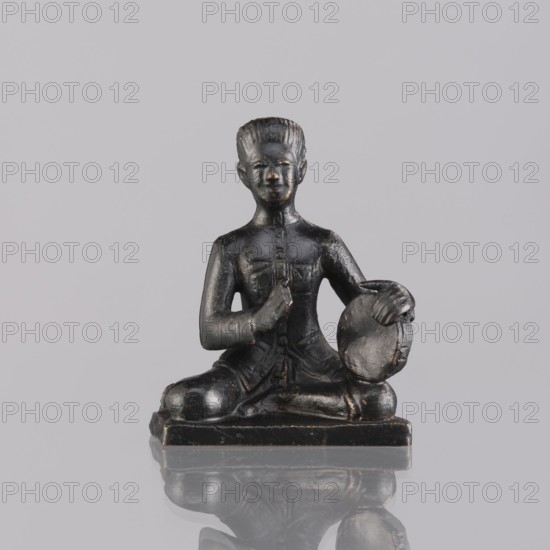Musician figurine, art of Southeast Asia, Southeast Asia culture, musicians, provenance unknown, 1st half of the 20th century contemporary product, Cambodia place of origin, sculpture, figurine, height 6.3 cm, A small dark statue of a seated figure is shown against a plain light gray background, the figure sitting cross-legged on a rectangular base with a reflective surface beneath that creates a faint mirror image; the statue appears predominantly black with subtle worn highlights of brownish-gold on raised areas, the figure has a stylized hairstyle, a defined face with neutral expression, and is holding a round object with one hand while the other hand rests near the chest, and the clothing and base show textured details and slight surface wear.