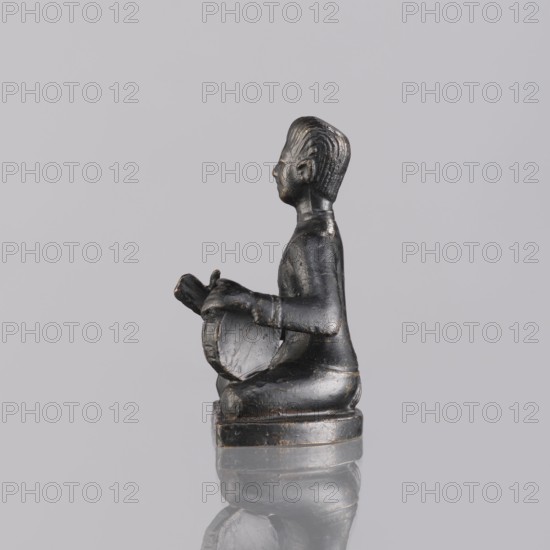 Musician figurine, art of Southeast Asia, Southeast Asia culture, musicians, 1st half of the 20th century contemporary product, Cambodia, sculpture, figurine, height 6.3 cm, A small dark figurine sits on a rounded base against a pale gray background, shown in profile facing left; the figure appears to be seated with bent legs and holds a rounded object in its lap with both hands, and the surface shows worn areas and slight variations of dark gray and black tones with some subtle lighter highlights.