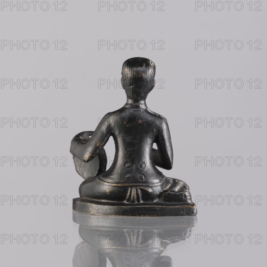 Musician figurine, art of Southeast Asia, Southeast Asia culture, musicians, provenance unknown, 1st half of the 20th century contemporary product, Cambodia place of origin, sculpture, figurine, height 6.3 cm, A small dark statue viewed from behind showing a seated human figure with short textured hair and a smooth back, arms bent with one arm resting on a rounded object, carved details on the lower back and waist, a draped cloth over the seated legs, and a flat base; the statue is primarily black with small areas of worn gold or brown showing, set against a light gray background and reflected faintly on the glossy surface beneath.
